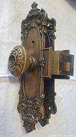 Side angle of one side of the ornate door hardware showing detailed metalwork on plate and knob, and the lock bolt extending from the mechanism.
