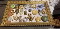 Top-down view of 28 costume jewelry brooches displayed on a rectangular gold-tone mirrored dresser tray, showing varied colors, shapes, and detailing.