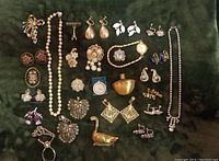 Overview of assorted costume jewelry including earrings, necklaces, brooches, cufflinks, rings, a small brass perfume bottle with glass insert, and a duck figurine on green velvet background.