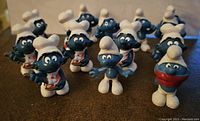 Fourteen blue Smurf figurines arranged on a surface, several dressed as chefs with white hats and aprons, one with red bandana, all appear in good condition.