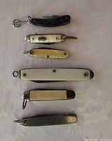 Photo showing six vintage miniature pocket knives laid out flat on a white surface.