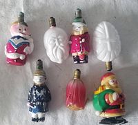 Top view of six vintage Christmas light bulbs showing various holiday-themed characters and shapes, including snowmen, Santa faces, and decorative bulbs.