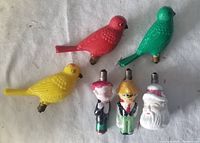 Photo shows six vintage Christmas light bulbs arranged on a white surface: three are colorful birds (red, green, yellow), and three are figural characters including Santa Claus and two other figures in painted glass.