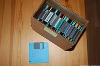 Box of approximately 40 used 3.5-inch floppy disks of assorted colors and labels on a wooden surface, one blue floppy disk placed in front of the box.