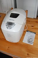 Full view of the white Black & Decker B6000C automatic bread maker showing the control panel and accompanying user manual placed on a wooden surface.