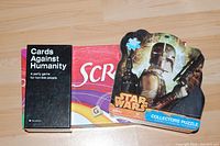 Photo of lot showing Cards Against Humanity (Canadian edition) in black box, Scrabble board game with classic purple box, and Star Wars Collector's puzzle with shaped box featuring The Mandalorian.