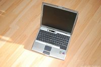 Dell Latitude D600 laptop open to show keyboard, touchpad, and blank screen on wood floor.
