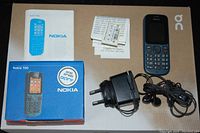 Photo of Nokia 100 phone next to its original box, user manual, charger, earphones, and paper ephemera on a table.