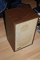 Full view of one speaker showing wood finish cabinet and fabric grille with faint stain