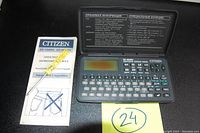 Photo showing the open Citizen ED-1500RX electronic organizer with LCD screen and bilingual keyboard, user manual, and metal stylus laid out next to the device.