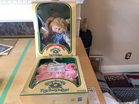 Front view of the boxed 1984 Cabbage Patch Kids doll alongside the boxed baby clothes set on table.