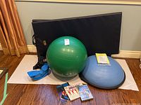 Full lot display showing green Boyce ball, blue Bosu balance trainer with yellow paperwork, blue plantar stretcher device, two black foam floor mats, and exercise booklets and DVD on wood floor.