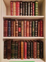 Three white bookshelf shelves filled with approximately 28 leather-bound hardcover books in various colors