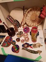 Full overview of the Indigenous folk art on a white table showing drums, carved mask, small baskets, painted folk art pieces, soapstone fish, and fur-covered animals.