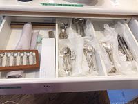 Drawer with multiple compartments holding various silverplate cutlery pieces wrapped in tissue paper, including teaspoons, forks, knives, and serving spoons.