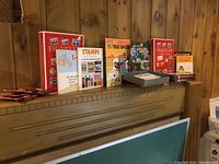 Photo of multiple stamp albums and books arranged on a wooden mantel, showing variety of books and albums related to stamps from Canada and around the world, including some loose stamps in an open box.