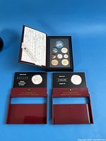 Three presentation cases open on blue background: one maroon case with uncirculated coins, two red cases labeled Proof Silver Dollar and Fine Silver $10