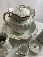 Group photo of the main pitcher with lid in the large matching basin and surrounding accessories showing gold gilt highlight details on cream porcelain.