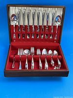 Opened wooden box with red velvet interior holding a set of silver plated flatware including knives, forks, spoons, and serving pieces, all with daffodil floral pattern on handles.