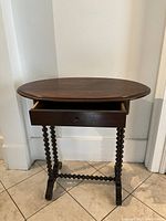 Oval walnut wood side table with open drawer and visible bobbin spiral legs on tiled floor against white wall.