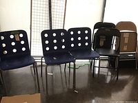 Photo showing three blue resin chairs with perforated backs and seats alongside three metal chairs (two folding, one more rigid). Reflective floor and grid metal cage in background.
