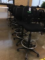 Row of eight black adjustable high office chairs with mesh backs, padded seats, caster wheels and chrome foot rings, shown on concrete floor indoors.