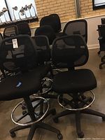 Eight black adjustable high office chairs with mesh backs, padded seats, footrings, and caster wheels grouped together indoors on a concrete floor.