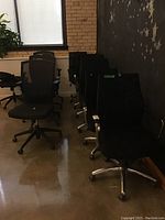 Photo showing a row of black office chairs with mesh backs and cushioned seats, lined up against a wall with a concrete floor reflecting light.