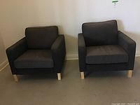 Two gray upholstered armchairs side by side against white wall, front view showing both chairs.