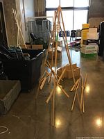 Several wooden tripod easels standing assembled on floor, with natural wood finish, adjustable center support and ledge for canvases