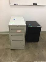 Photo showing two metal filing cabinets side by side, one white with three drawers and one black with two drawers, set in a room with white walls.