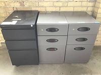 Three metal filing cabinets side-by-side against a brick wall: one black with three drawers, two gray with two drawers each, showing wear.