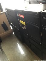 Side view of six black two-drawer metal filing cabinets arranged in a row showing the drawer handles and sticker details.