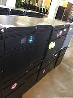 Photo showing a group of black metal filing cabinets arranged side by side, each with two drawers and stickers on front panels.