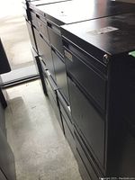 Side view of multiple black metal filing cabinets lined up in a row, showing two drawers each and lock cylinders without keys.