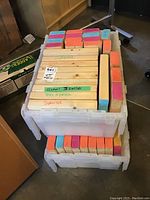 Two large plastic containers packed with large-sized Jenga wooden blocks, some painted in pastel colors and some natural wood with printed labels.