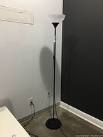 Full view of the standing floor lamp positioned in a corner with visible base, pole, and white upward-facing shade against a two-tone wall.