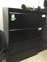 Full view of the black metal 4-drawer filing cabinet shown from the front and side angle, with a chair placed beside it.