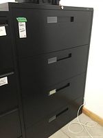 Front view of the black metal filing cabinet showing all 4 drawers and lock area on top drawer, display tags on the left side of the cabinet.