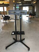 Full view of the adjustable metal monitor stand showing structure, wheels, and mounting bracket.