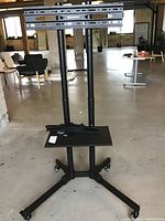 Full view of the black metal adjustable monitor stand with wheels and mounting bracket in an industrial space.