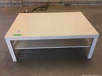 White rectangular melamine coffee table with a second shelf underneath, standing on a concrete floor against a brick wall, visible wear on surface.