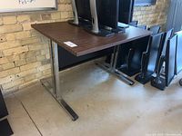 View of the table from an angle showing the wood laminate top and metal legs with chrome finish, on a concrete floor against a brick wall.