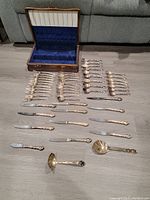 Full layout of 50-piece Oneida silver plated flatware set with storage chest open behind, showing polished spoons, forks, knives, and serving utensils on floor.