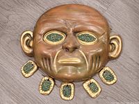 Front view of the ceramic mask showing the wrinkled face design, golden accents, green textured inset eyes, and surrounding dangle elements.