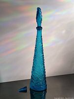 Front view of tall blue Empoli genie bottle with broken stopper placed beside it on surface, showing tapered shape and textured wave pattern.