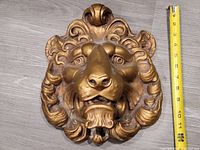 Front view of a gold-colored plaster lion head showing detailed mane and face with measuring tape for scale (approx. 12" tall).