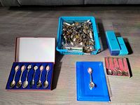 Photo showing a large blue plastic basket filled with assorted loose collector spoons, a boxed set of six spoons with decorative oval motif handles in a blue case, a single spoon in a blue plastic case, and a smaller set of four ornate spoons in a red-lined box.