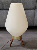 Front view of the beehive lamp showing the ribbed off-white shade, brass fitting, and teak tripod legs sitting on a wood floor in front of a sofa.