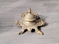 Brass inkwell displayed closed on a wood floor showing ornate patterns on the lid and base.